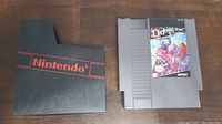 Front view of Defender of the Crown NES cartridge with colorful game label next to black Nintendo sleeve with red logo.