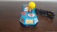 Front left angled view showing yellow joystick, two red buttons, NAMCO brand logo with game art stickers on angled sides, and attached AV cable.