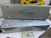 Photo showing two boxed Forever scooters stacked, silver boxes with black text and graphics, and a US dollar bill for size reference.