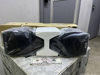 Front view of two KLH HT-200 speakers, wrapped in plastic, placed on styrofoam, sitting on top of original box, showing size reference with $5 bill.