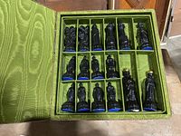 Top view of the fabric-covered box open showing black ceramic chess pieces organized in individual slots on a green fabric interior.