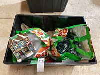 Photo of the black plastic tote containing the lighted display house, two bags with ornaments, and string of lights. The bag on the left shows the front of one ceramic display house with a wreath and festive ribbons, along with wrapped ornaments.