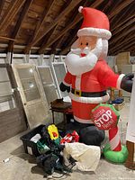 Large inflatable Santa Claus holding a 'Santa Stop Here!' sign pictured indoors in attic-like space, with plastic tote and other items on the floor.
