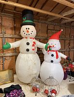 Two snowman Christmas inflatables side by side, large one with a top hat and smaller one with a Santa hat, surrounded by Christmas decorations and storage tote