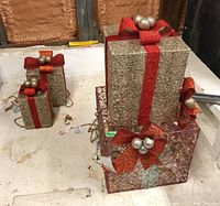 Four lighted outdoor Christmas gift boxes displayed on the floor with visible plugs and cords.