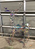 Frontal view of metal wire frame deer with iridescent mosaic panels and string lights against garage door backdrop.