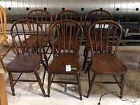 Six wooden dining chairs arranged in two rows on concrete floor, showing overall appearance, wood finish, spindle backs, and wear.