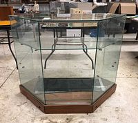 Front view of angled glass and veneer display cabinet showing clear glass sides, wooden base, and visible wear on veneer.