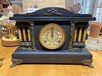 Front view of black wood mantle clock with green faux marble columns, ornate gold clock face with Arabic numerals.
