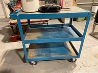 Right side view of the blue metal cart with three shelves and caster wheels, showing the overall structure and dirt on shelves.