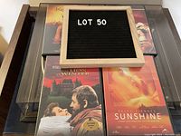 Photo of three DVDs displayed on a glass table showing 'The Lion in Winter', 'Kundun', and 'Sunshine' DVDs with a sign indicating Lot 50.