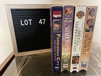 Four VHS tapes showing the spines with titles Forbidden City, Shadows, The Bridges of Madison County, and Sense and Sensibility arranged vertically next to a letter board labeled Lot 47.