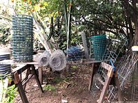Photo showing roll of green wire mesh fencing, coiled rolled wire mesh, blue edging stakes, and a metal trellis on a wooden bench in garden setting under orange tree.