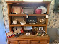 Wide shot of wooden shelf with kitchenware items arranged including stoneware dishes, bakeware, glass dishes, salt and pepper shakers, iron warmers, napkin rings and magnet collection.