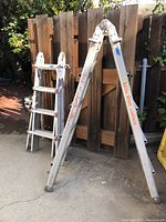 Two ladders set up next to fence: 17 ft multi-position ladder open and 10 ft ladder folded.