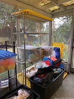 Full view of metal shelving unit with four wire mesh shelves, some storage bins and items on it, top shelf with yellow cover, unit on wheels in a screened porch area.