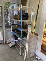 Wide photo showing taller six-shelf assembled white metal wire shelving unit with miscellaneous items on shelves and second shorter shelving unit disassembled to right side.