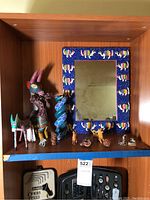 Full view of shelf containing multiple colorful hand-carved Mexican Alebrijes, smaller Asian wooden figurines, small ceramics, and a wall mirror with Alebrijes painted frame.