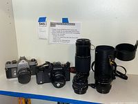 Photo showing two film SLR cameras with multiple lenses and accessories arranged side by side, with placard listing included items.