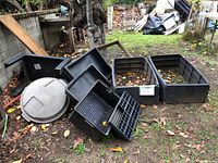Photo of black plastic raised beds, seeding trays, bin lids, and other items arranged on ground with fallen leaves and debris.