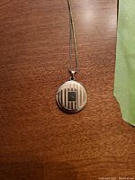 Front view of round 14k gold locket with stripes and small square detail on front, hanging from chain on wooden surface.