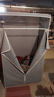 Gray vinyl storage unit with zippered opening partially open revealing blankets and pillows inside. Shows front view with transparent top section and metal rack inside.
