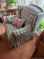 Front view of wingback chair showing green and cream plaid upholstery with floral cushion on the seat, and wooden frame at the top backrest.