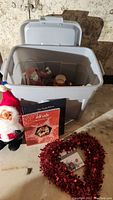 Overall photo of plastic storage bin containing various Christmas decorations and books around it, including a red metallic heart-shaped wreath and stuffed Santa doll.