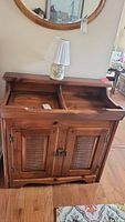 Front view of vintage wooden dry sink with two open slanted compartments on top, dual louvered door cabinet below, and lamp on top in a room setting.