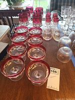 Front view of 18 pieces of glassware, including cranberry rimmed and clear glasses grouped on a table.