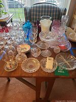 Wide view of entire assortment of glassware showed on table in front of window with plaid chair background, includes blue glass bottles, clear wine glasses, bowls, and vases.