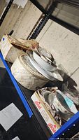 Photo of assorted china pieces stacked on shelf, showing cups, plates, bowls, some wrapped in newspaper and packed carefully in boxes.
