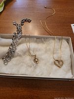Three necklaces laid out in a box lined with white fabric for display: silver-tone linked ring chain, gold-tone heart pendant with opal-like stone, gold-tone heart outline pendant with stones.