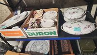Photo showing several Limoges dinnerware pieces including a stack of plates, candy dish, and covered serving bowls with floral patterns and gold trim on a basement shelf.