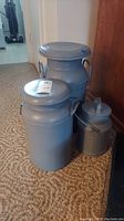 Three metal painted milk containers arranged on carpet floor beside wooden furniture.