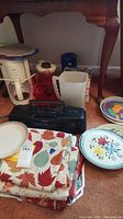 Photo showing white Krups coffee maker with glass carafe and several plastic pitchers, a couple of decorated plates, and folded table linens with leaf designs.