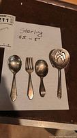 Four small sterling silver flatware pieces laid out including two spoons, a fork, and a pierced dessert spoon.