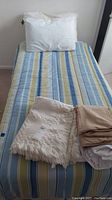 Twin bed made with blue, green, and white striped bedspread, pillow with white embroidered pillowcase, beige sheets, and fringed blanket on top.
