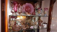 Glass cabinet shelf with midcentury cranberry glass pitchers, vases, and ruffled bowls, including Depression glass stemware and candy dishes. Milk glass floral vases and porcelains also visible.