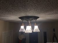 Photo of four-light vintage ceiling fixture mounted on a textured ceiling, showing all four frosted glass bell-shaped shades with leaf etching and dark metal ornate base.