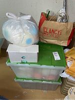 Image showing plastic bins with green lids stacked containing yarn and supplies, plus paper bags and a plastic bag on top.