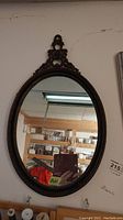 Full view of the wood framed oval wall mirror with ornate carved detail at the top, hanging on the wall in a basement space.