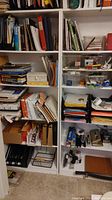 Wide view showing large white shelves filled with books, folders, papers, and office accessories including stapler, hole punch, and a paper cutter.