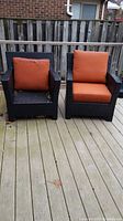 Two wicker outdoor arm chairs on a wooden deck, one with both orange cushions and one with seat cushion only