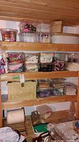 Four wooden shelves in basement holding multiple plastic containers and boxes filled with fabric pieces, lace, cords, and other sewing notions.