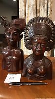 Front view of two wooden Balinese warrior busts with visible detailed carvings and dark wood finish, along with pen and lot tag for scale.