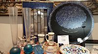 Wide shot of art glass items on display including blue glass vases, stained glass panel, constellation plate, and mosaic plate with ceramic jugs in background