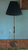 Full view of the vintage Danish floor lamp with a tall slender brass pole, green pleated lampshade and round brass base standing on a wooden floor.