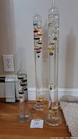 Full view of three vintage Galileo thermometers in various heights showing colored floating bulbs inside clear glass cylinders.