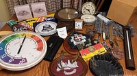 Wide shot showing multiple items on a wooden table including clocks, metal signs, wooden plaques, and other collectibles distributed across the surface.
